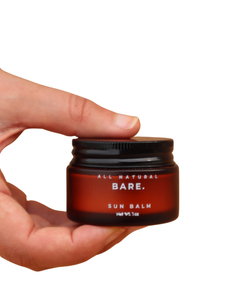 Hand holding a red and black container of Bare All Natural Sun Balm on a white background